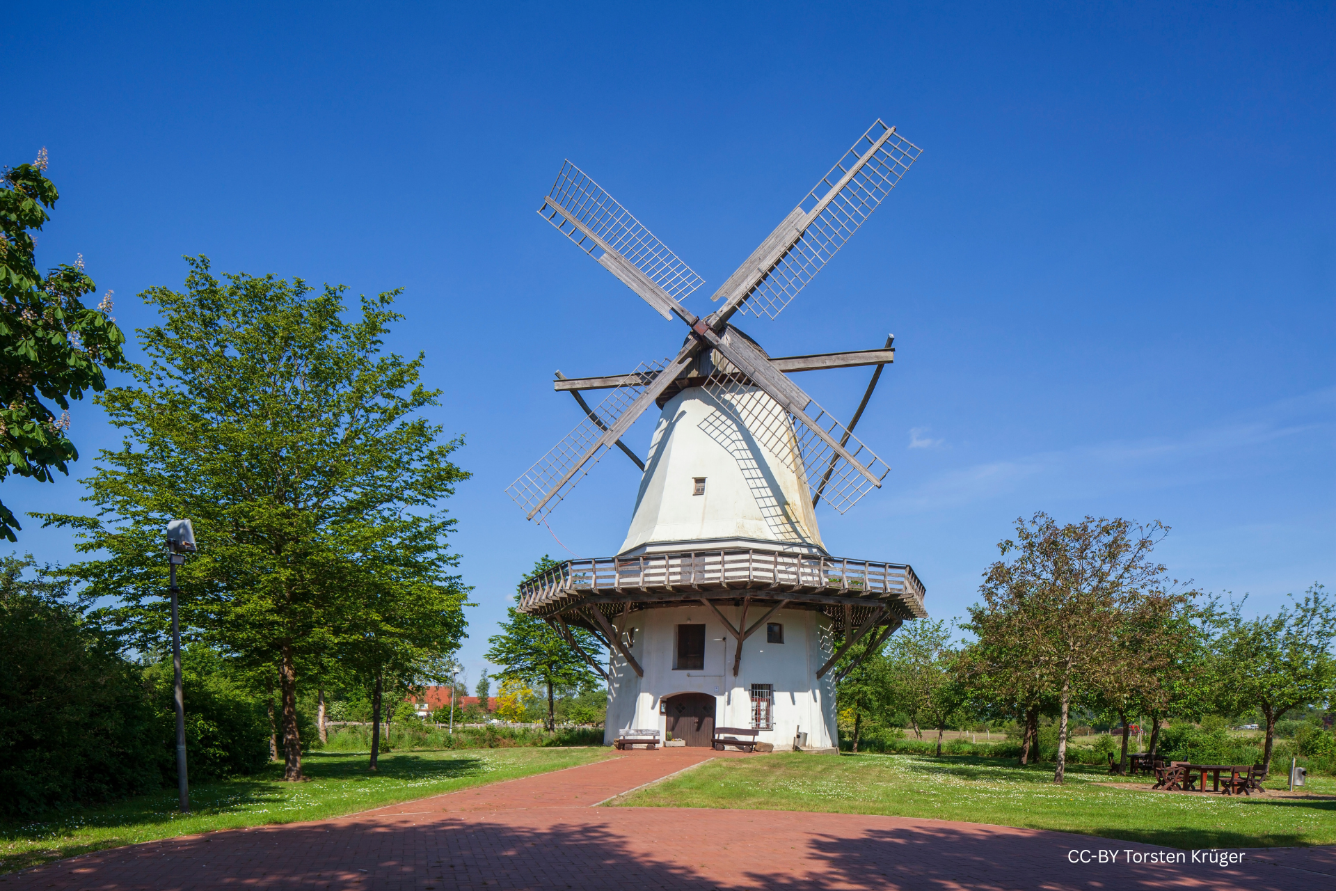 Historische Windmühle in Tonnenheide mit umlaufendem Balkon, rotem Ziegelweg und umgebender Grünfläche unter blauem Himmel.