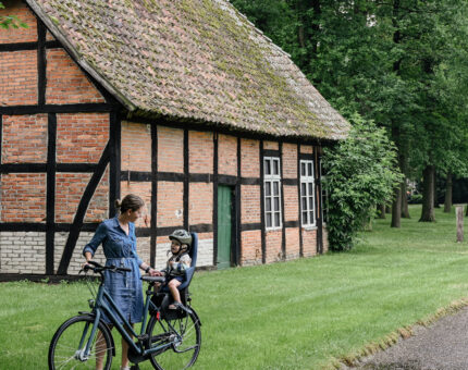 Eine Frau steht vor einem Fachwerkhaus mit einem Fahrrad, auf dem im Kindersitz ein Kleinkind sitzt.