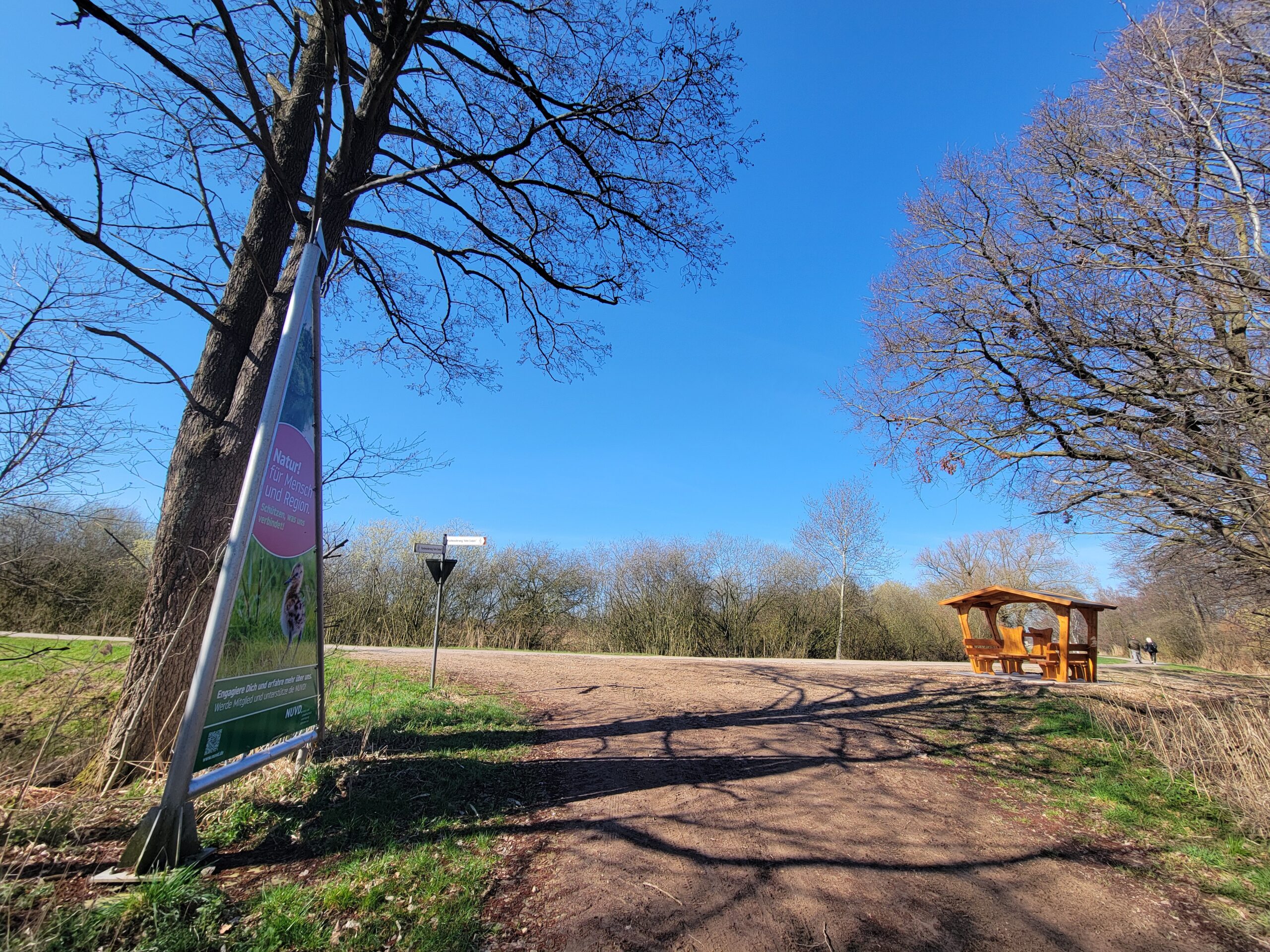 unbefestigter Wanderweg mit einem dreieckigen bedruckten Segel am linken Wegrand und einem überdachten Picknickplatz am rechten Wegrand