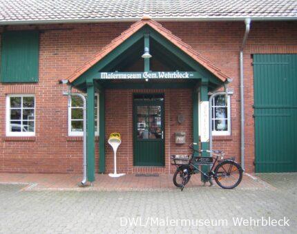 Eingangsbereich des Malermuseums Wehrbleck mit rotem Klinker, grünen Holzelementen und einem klassischen Malerfahrrad vor der Tür.