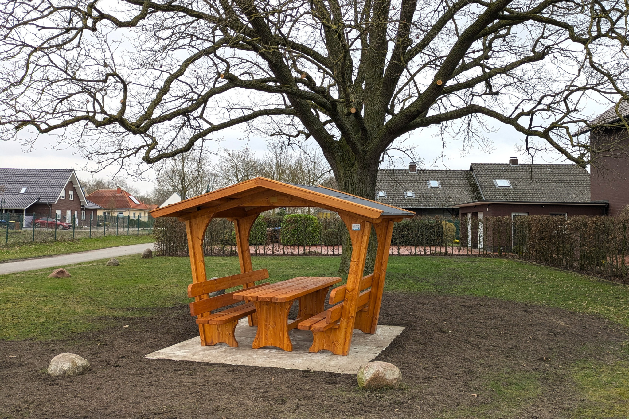 Ein überdachter Picknickplatz aus Holz mit zwei Bänken und einem Tisch auf einem grasbewachsenen Grundstück mit einer großen Eiche im Hintergrund