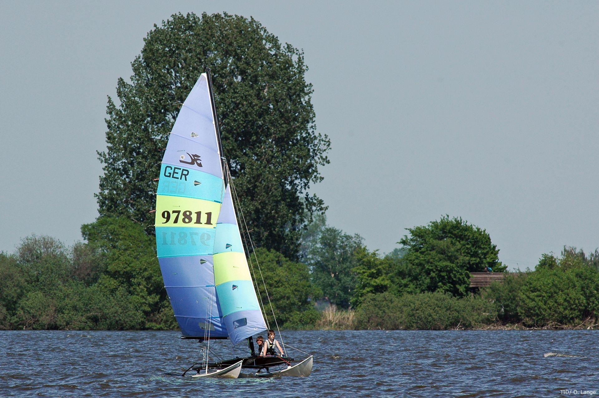 Zwei Personen segeln auf einem sportlichen Katamaran mit buntem Segel über den Dümmer, im Hintergrund dichter Baumbestand.